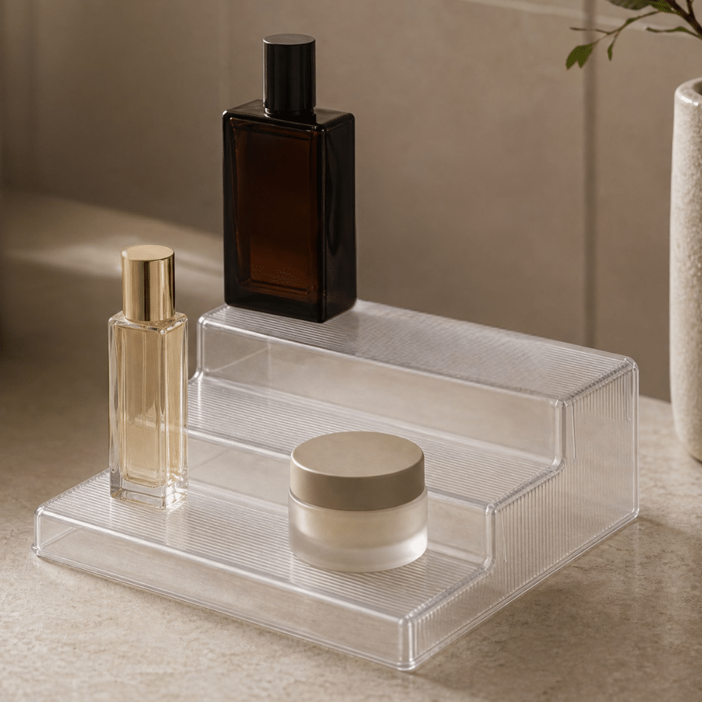 organisering på badeværelset med et plast podium | A clear acrylic tiered organizer holding two bottles and a small round jar on a beige countertop.