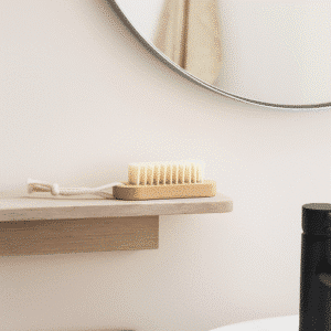 Lille træneglebørste med hvide børstehår ligger på en lys trähylde i et minimalistisk badeværelse foran et rundt spejl og en sort armatur.