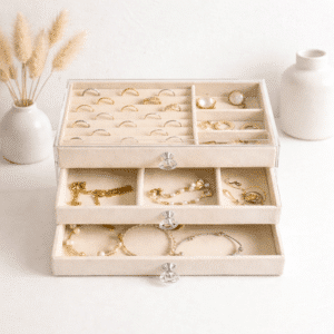 Smykkeskrin i klar akryl med tre udtrækkelige skuffer og beige stofindtræk til ringe, øreringe, halskæder og armbånd, organiseret og beskyttet.