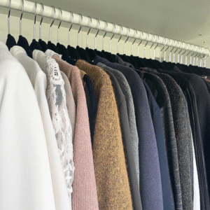 Rows of coats and jackets hanging on a metal clothes rack in a shop or closet, showing white, pink, brown, gray, navy, and black garments side by side.